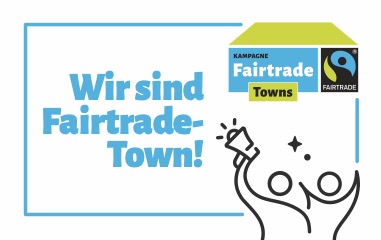 Die Grafik stellt das Fairtrade Logo dar und beinhaltet in blauer Schrift den Text: "Wir sind Fairtrade-Town".